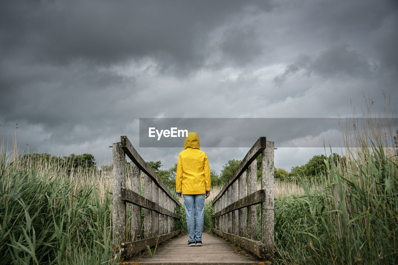 Rear view of a person standing in the middle of a wooden bridge with a dark moody sky