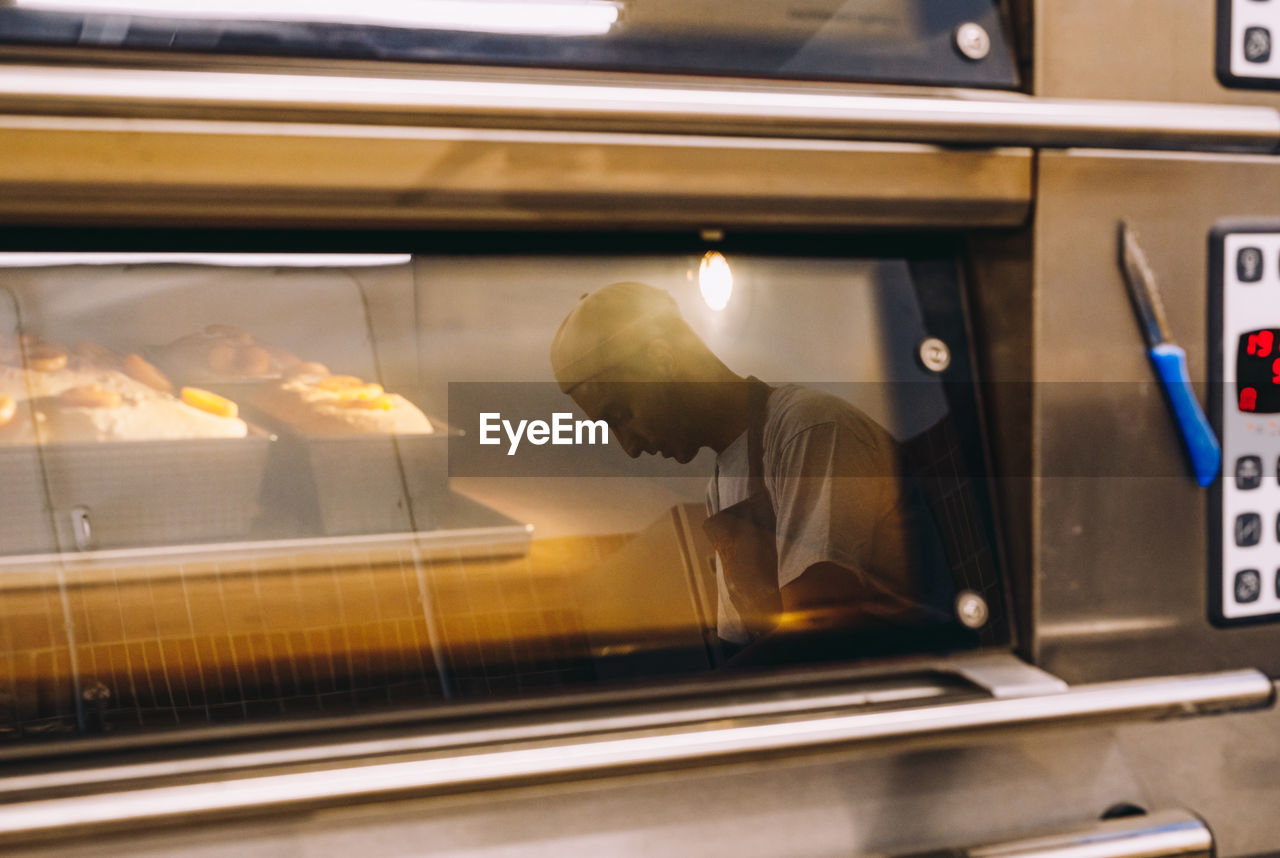 Side view of male baker in uniform standing in kitchen and reflected in glass of oven with tasty bread