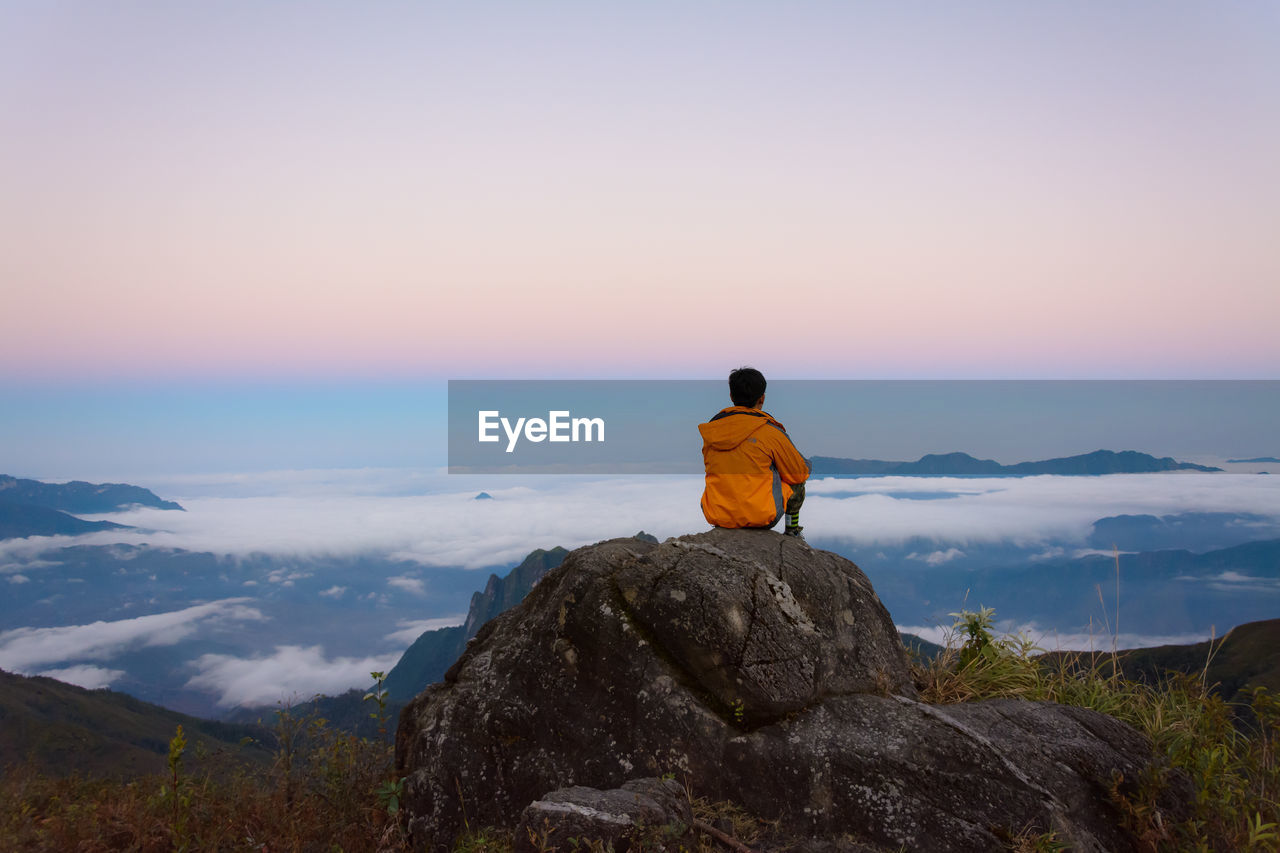 Rear view of man sitting on rock against sky