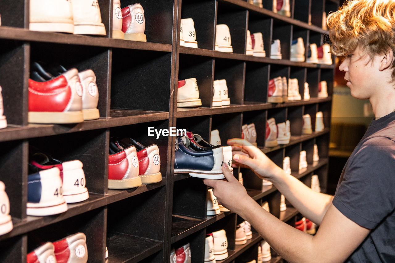 Side view of blond teenage boy holding bowling shoes at wooden rack