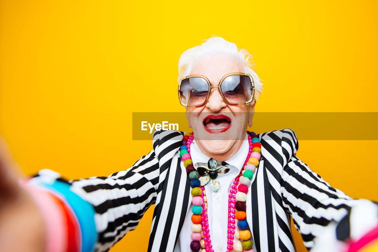 Portrait of stylish senior woman wearing colorful jewelry standing against yellow background