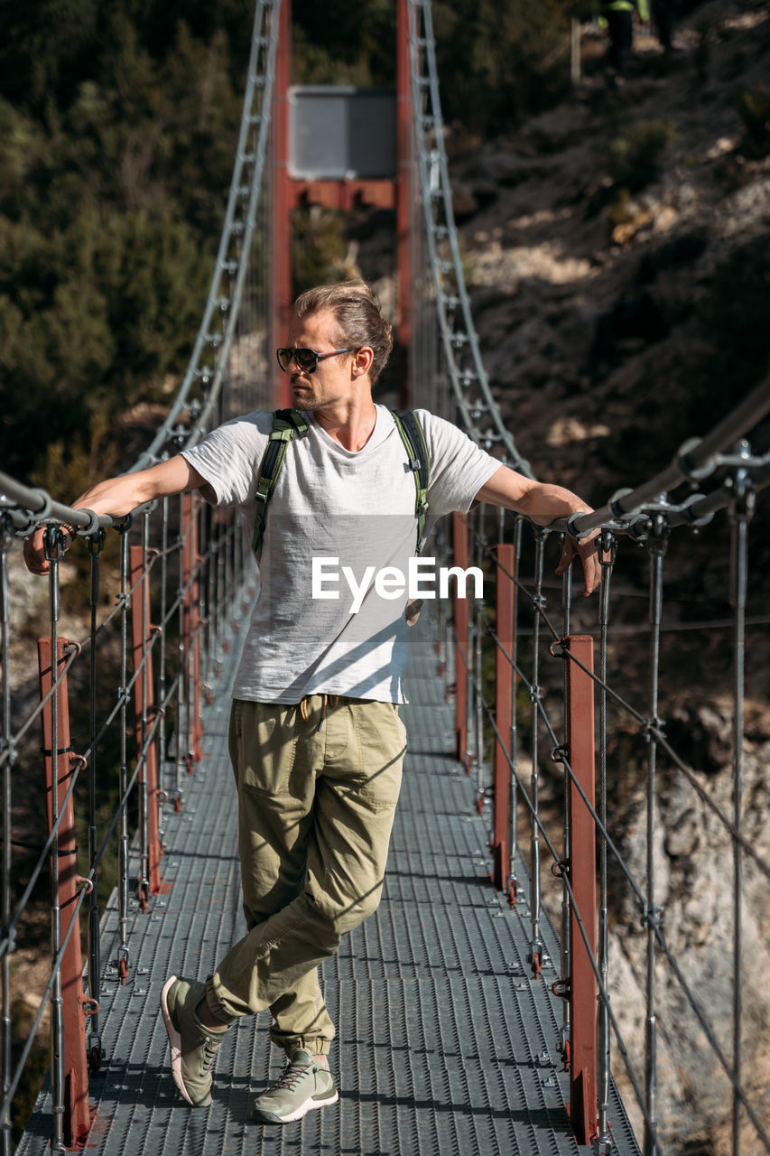 Man hiking in the mountains with backpack, standing on the bridge on sunny day.