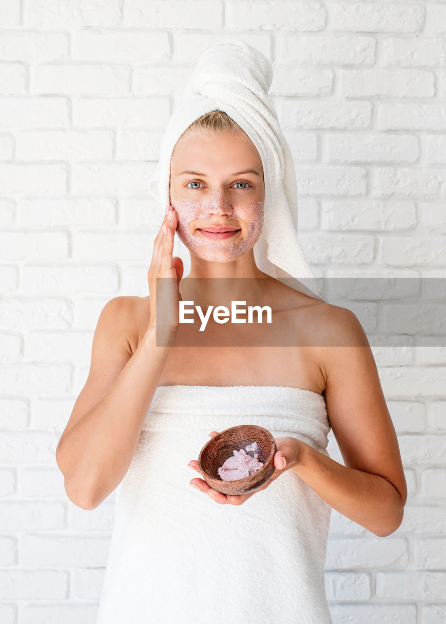 Young woman wearing white bath towels on head doing spa procedures applying facial scrub