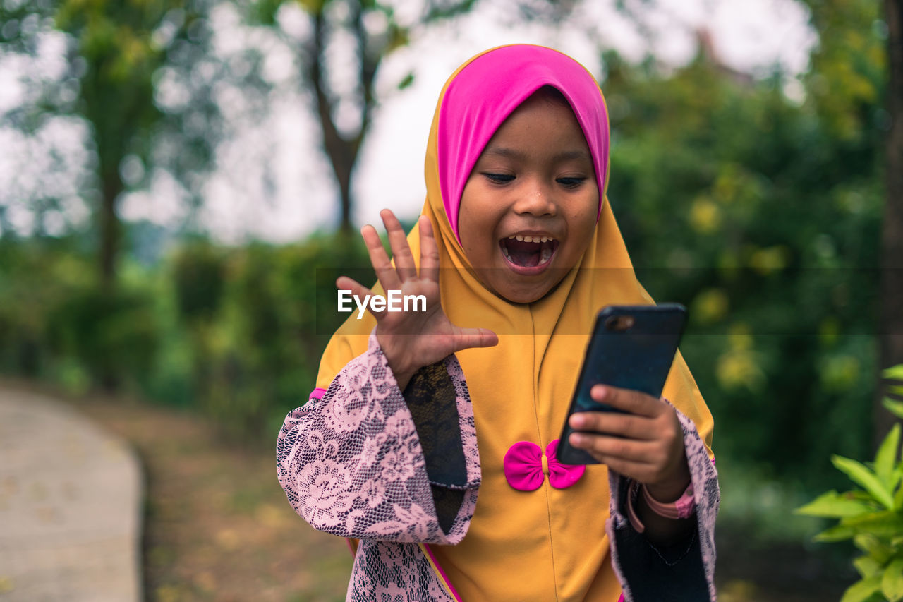 Cheerful girl wearing hijab using smart phone while standing at park 