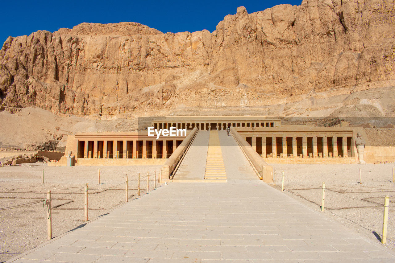 architecture, wadi, ancient history, travel destinations, desert, built structure, travel, history, nature, ancient, the past, no people, sky, sand, tourism, land, scenics - nature, mountain, temple, landscape, religion, ancient civilization, environment, sunny, rock, monument, clear sky, outdoors, arid climate, arch, historic site, day, sunlight, climate, natural environment, rock formation, building exterior, empty, blue, valley, landmark, the way forward, belief, archaeology, temple - building
