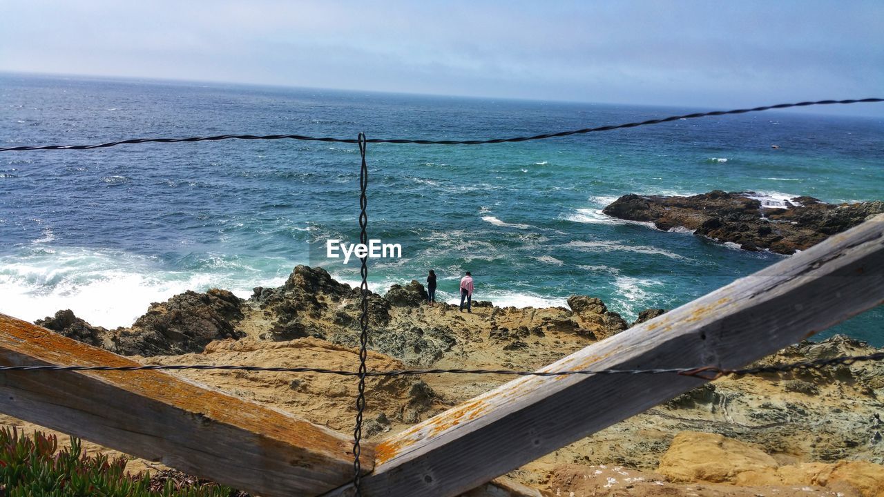 Disruptive view through wire fence of couple standing on ocean cliff. 
