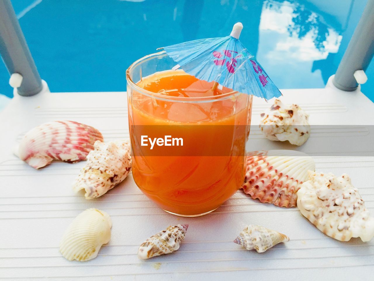 Glass of orange juice with blue umbrella and seashells near the blue pool water