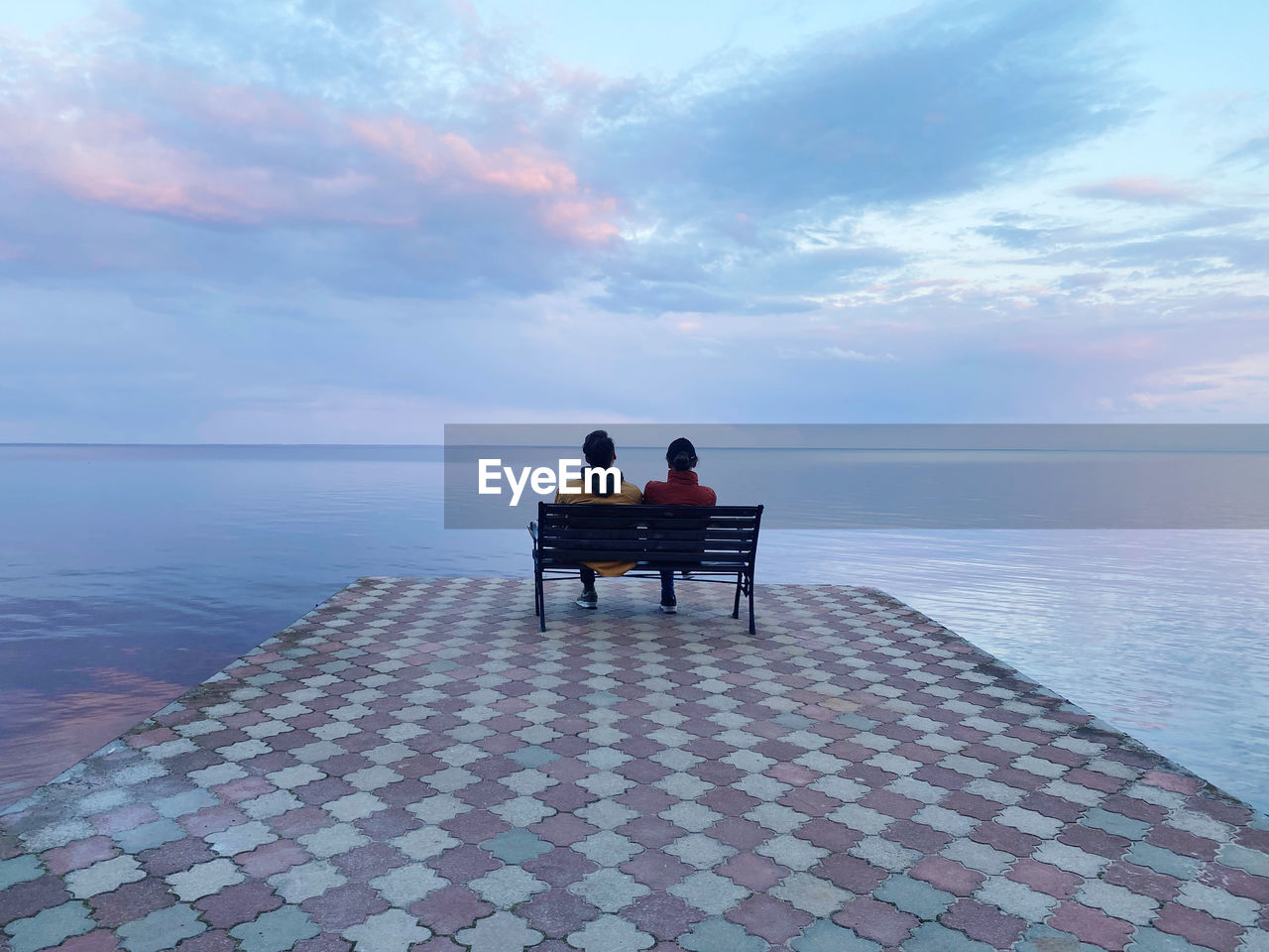 Rear view of men sitting on bench looking at sea against sky