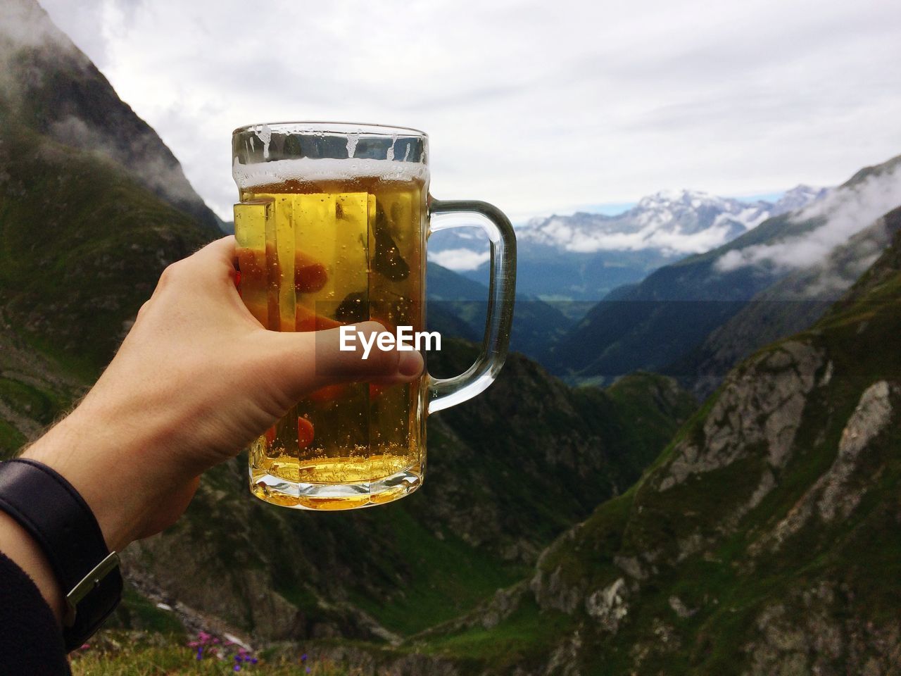 CLOSE-UP OF HAND HOLDING BEER GLASS AGAINST MOUNTAIN RANGE AND SKY