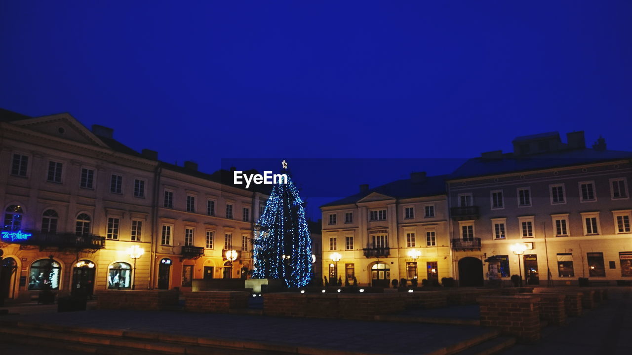 Christmas tree in front of illuminated buildings against clear blue sky