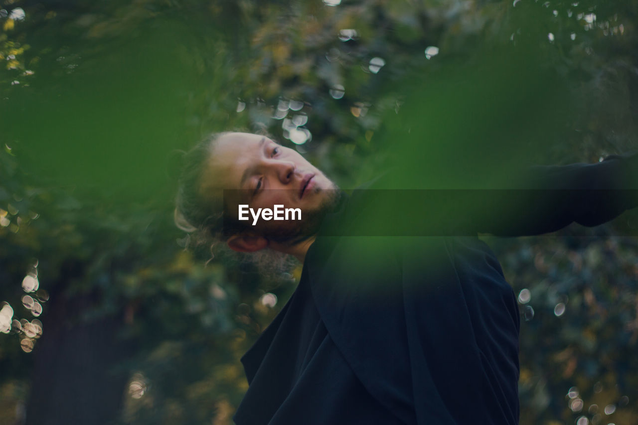 Thoughtful young man looking away while gesturing while standing against trees 