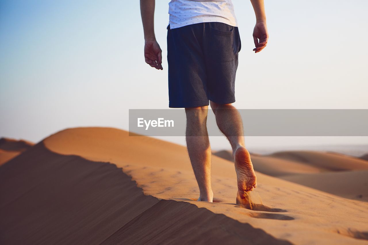 Low section of mid adult man walking on sand at desert against clear sky during sunset