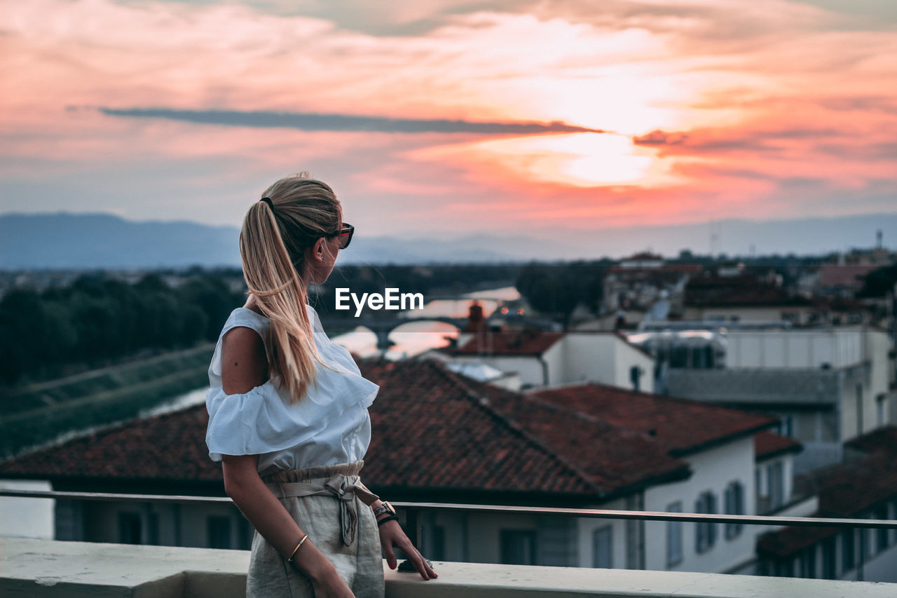Woman looking at cityscape against sky during sunset