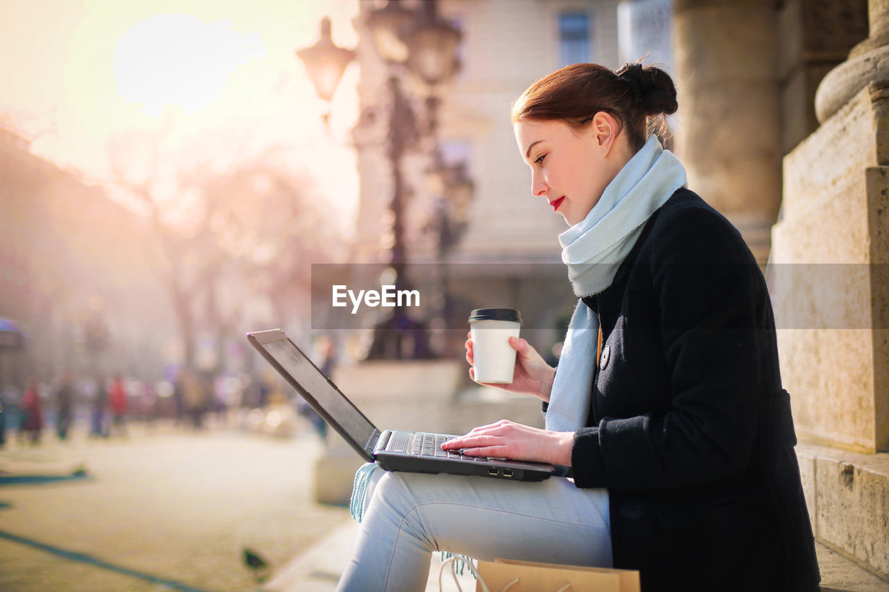 Woman using laptop while sitting on bench in city