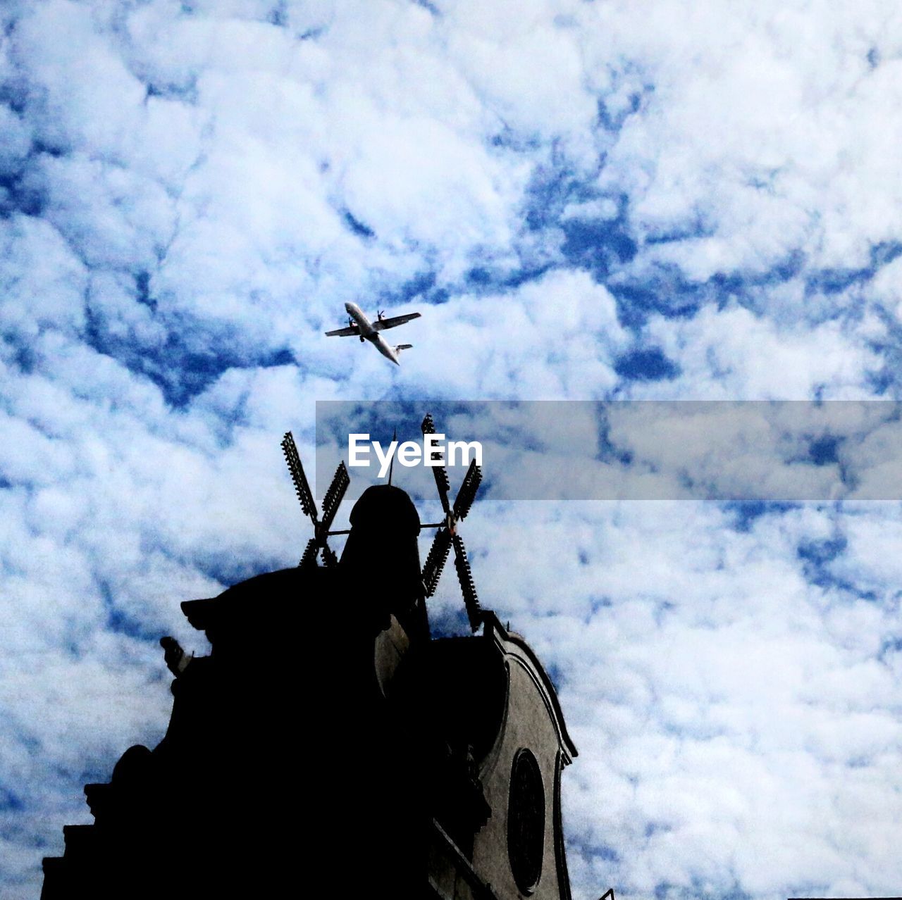 Airplane flying over traditional windmill against cloudy sky