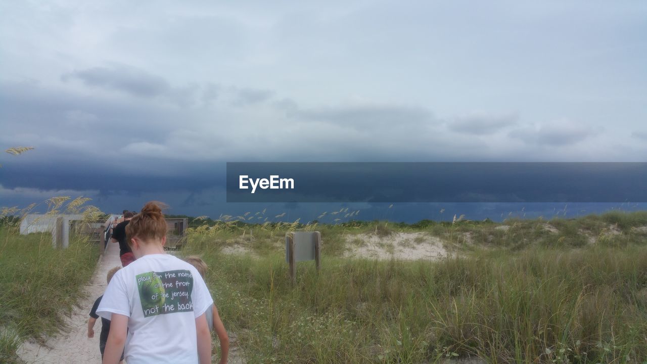 REAR VIEW OF WOMAN STANDING ON GRASSY FIELD AGAINST CLOUDY SKY