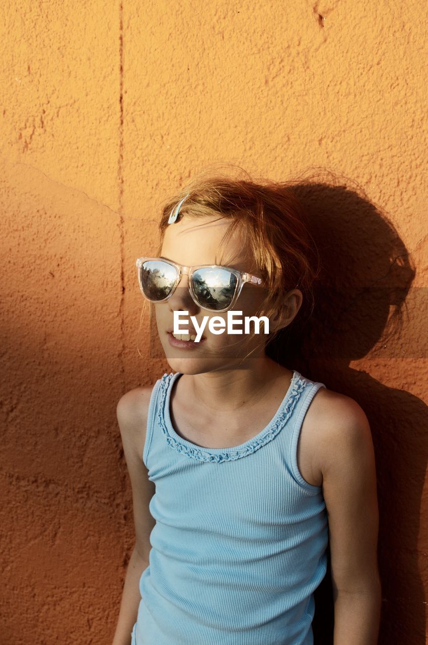 Portrait of young girl wearing sunglasses standing against wall