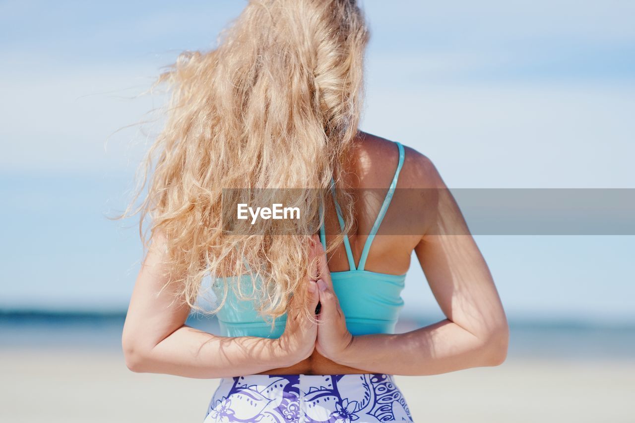 Girl standing at the beach in a yoga pose, facing the sea with wind blowing her hair