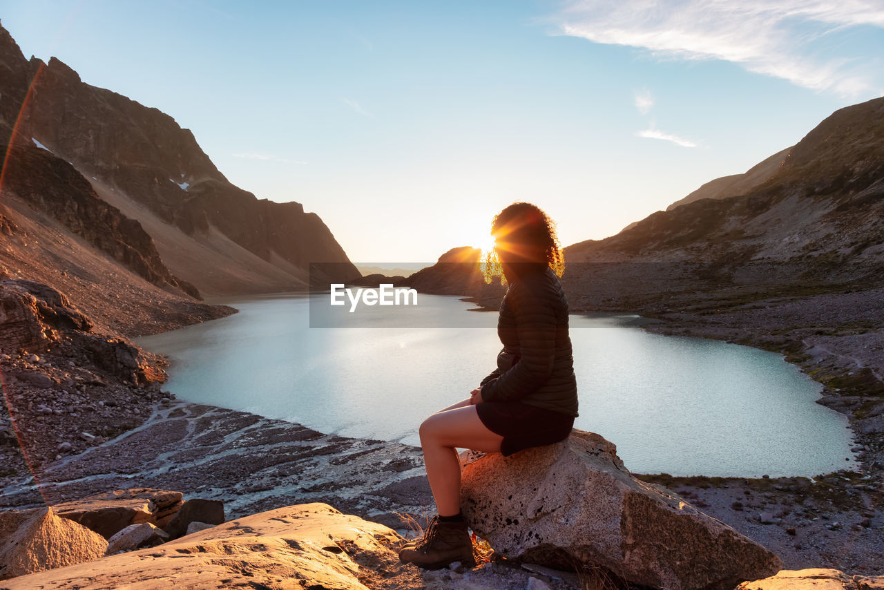 MAN SITTING ON ROCK AT SHORE AGAINST SKY