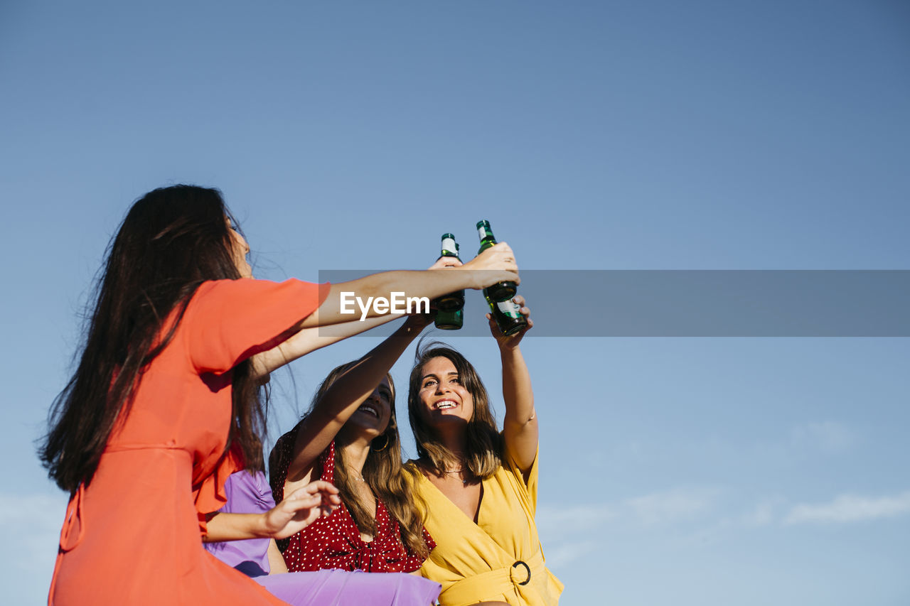 From below of group of joyful female friends toasting and clinking glass bottles of beer