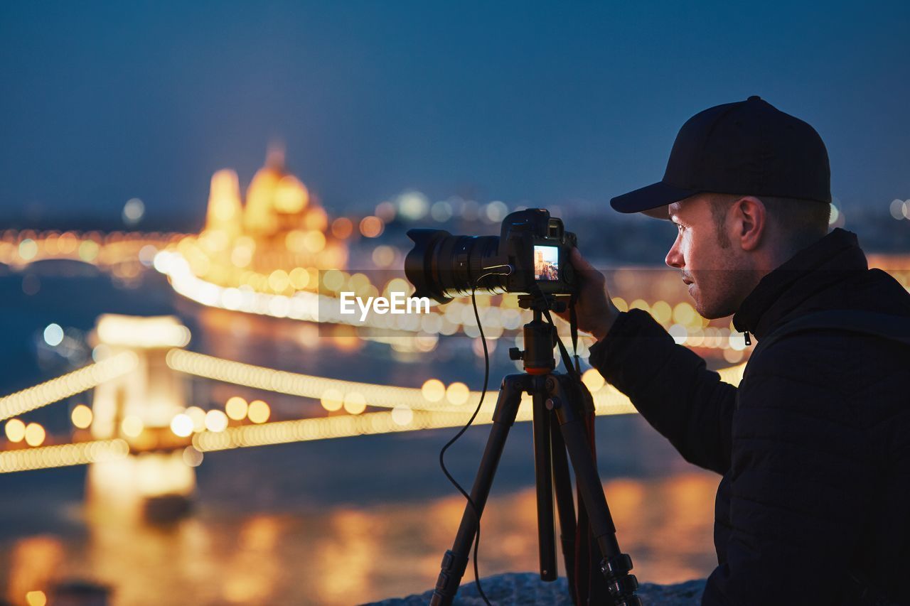 Man photographing illuminated chain bridge from camera in city at night