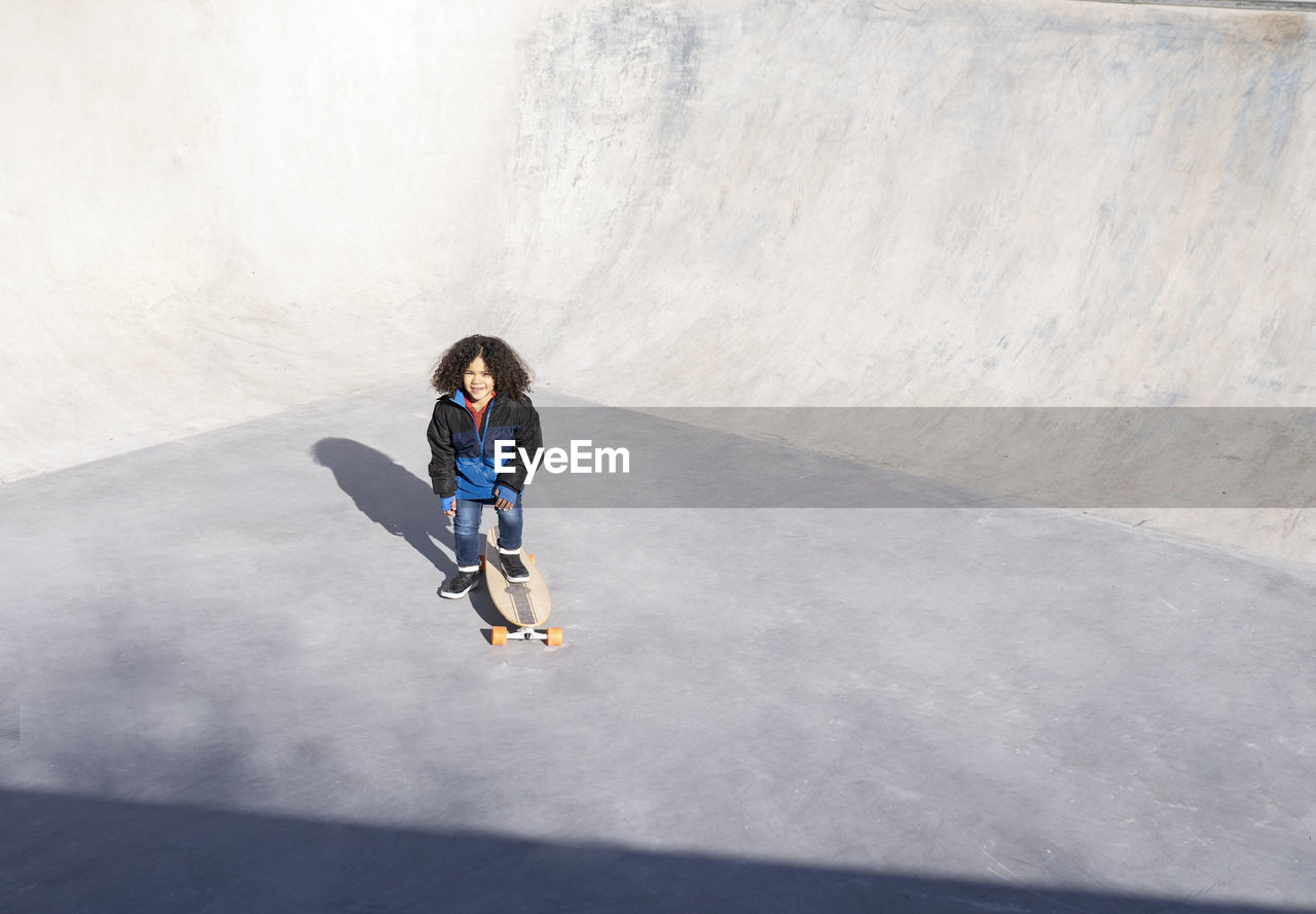 Ethnic child with curly hair standing on ramp with longboard in skate park and looking at camera