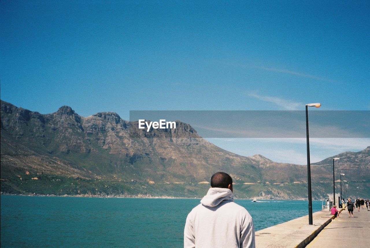 REAR VIEW OF MAN LOOKING AT SEA AGAINST MOUNTAIN
