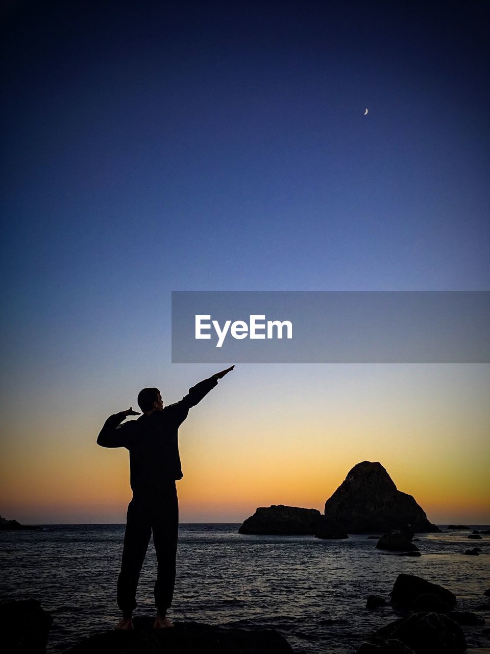 Rear view of man gesturing at sea shore against sky during sunset