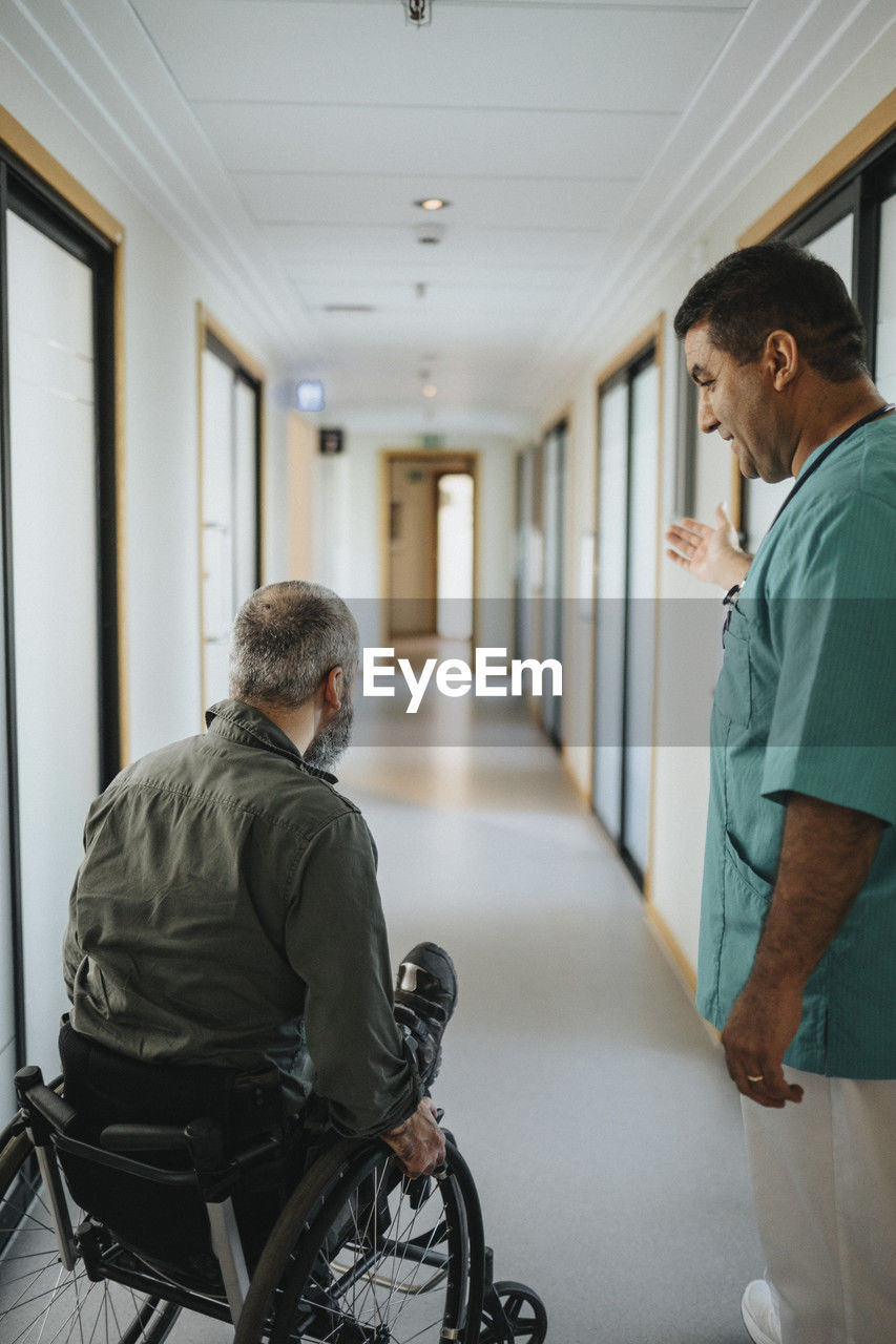 Male doctor showing way to male patient sitting in wheelchair at hospital corridor