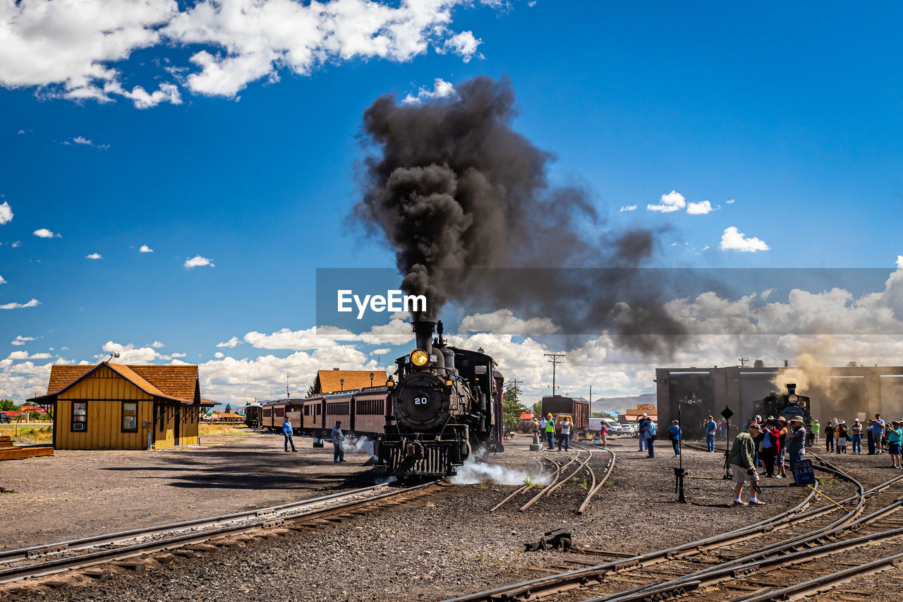 train, smoke, rail transportation, sky, railroad track, railway, transport, track, vehicle, locomotive, cloud, transportation, architecture, nature, mode of transportation, land vehicle, steam train, building exterior, steam, motion, built structure, group of people, public transportation, outdoors, day, pollution, blue