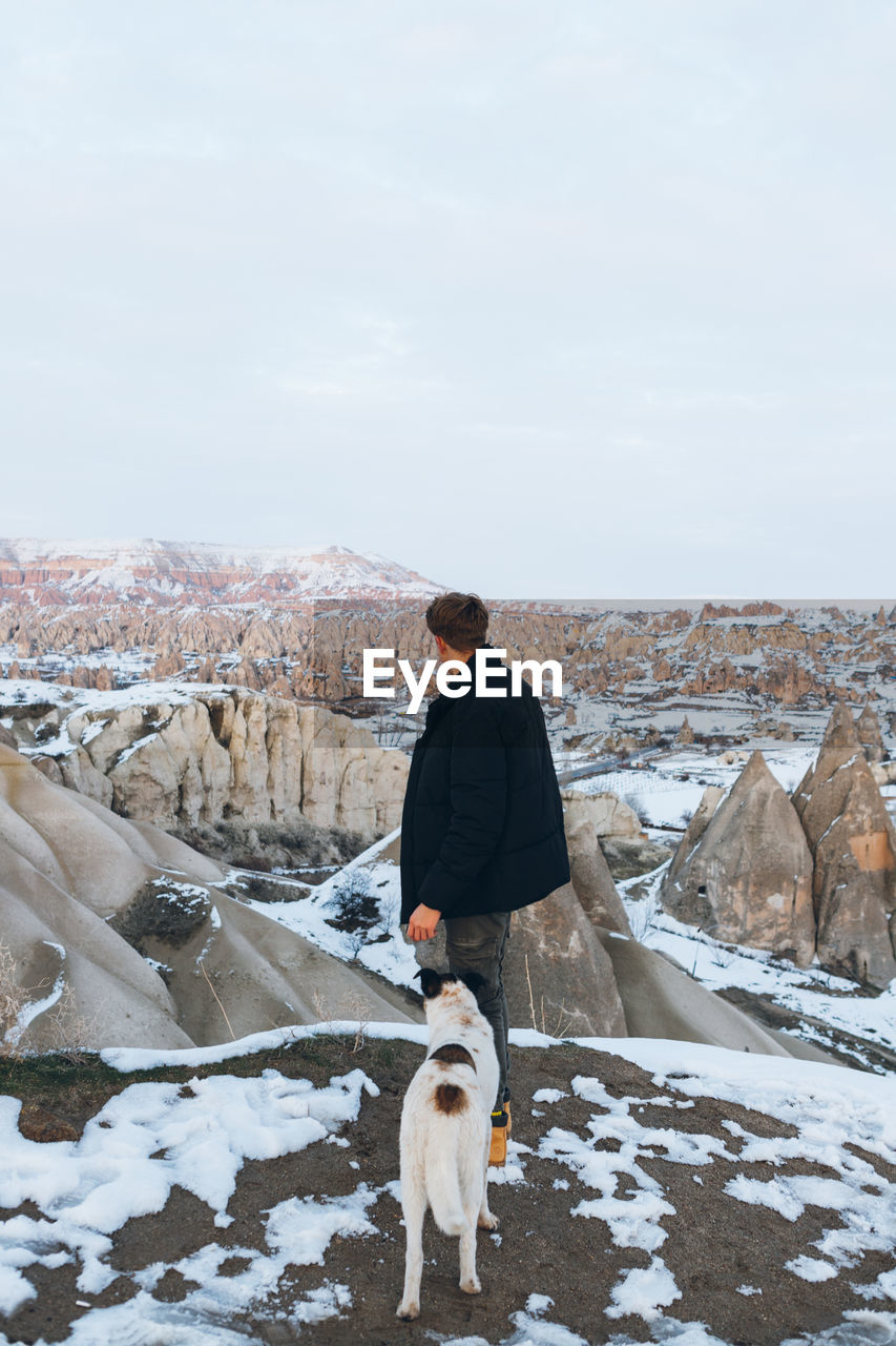 Back view of unrecognizable man in warm clothes with loyal dog in snowy hill against misty mountains in overcast weather in turkey