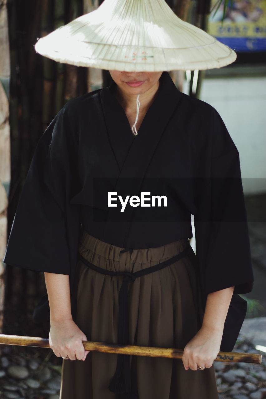 MIDSECTION OF WOMAN WITH HAT STANDING AGAINST BLURRED BACKGROUND