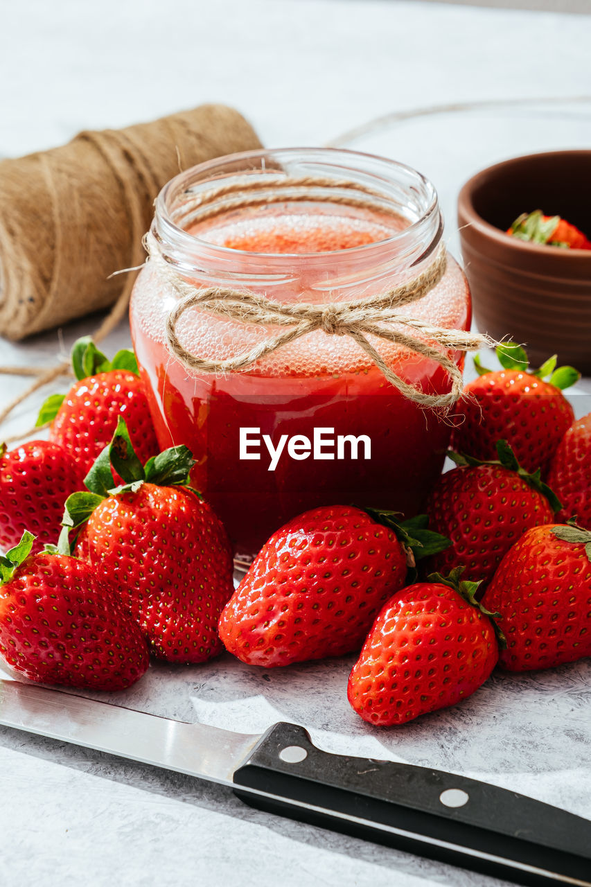 Composition with fresh homemade strawberry juice in glass jar wrapped with twine placed on marble surface with whole berries and knife