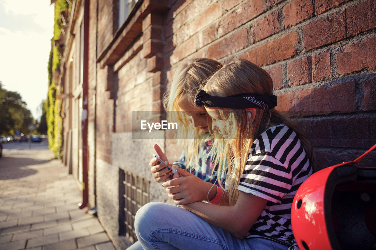 Girls using mobile phone while sitting against brick wall on sunny day