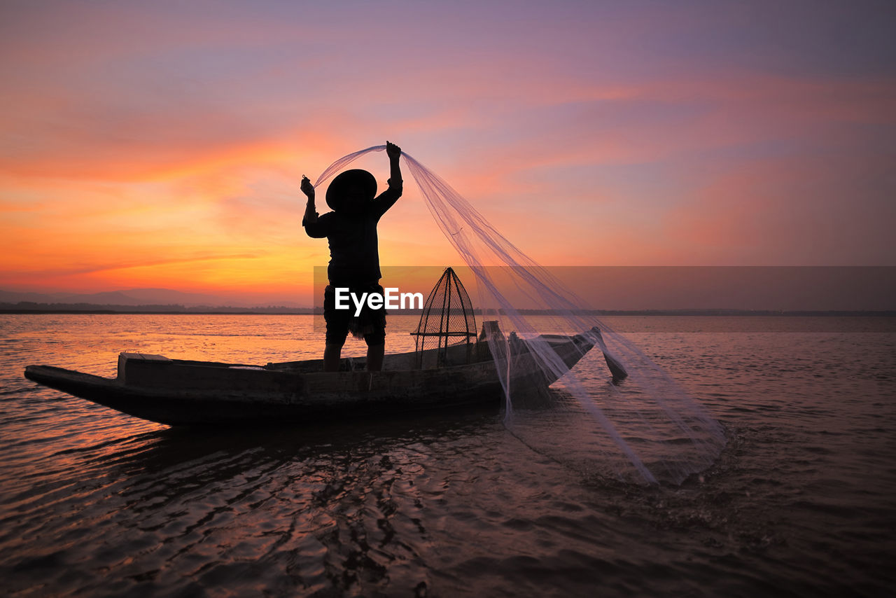 Silhouette fisherman holding fishing net in boat on sea during sunset