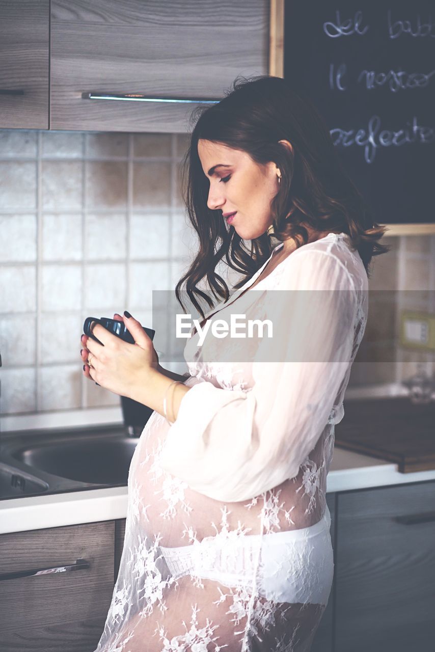 Side view of pregnant young woman holding mug while standing in kitchen