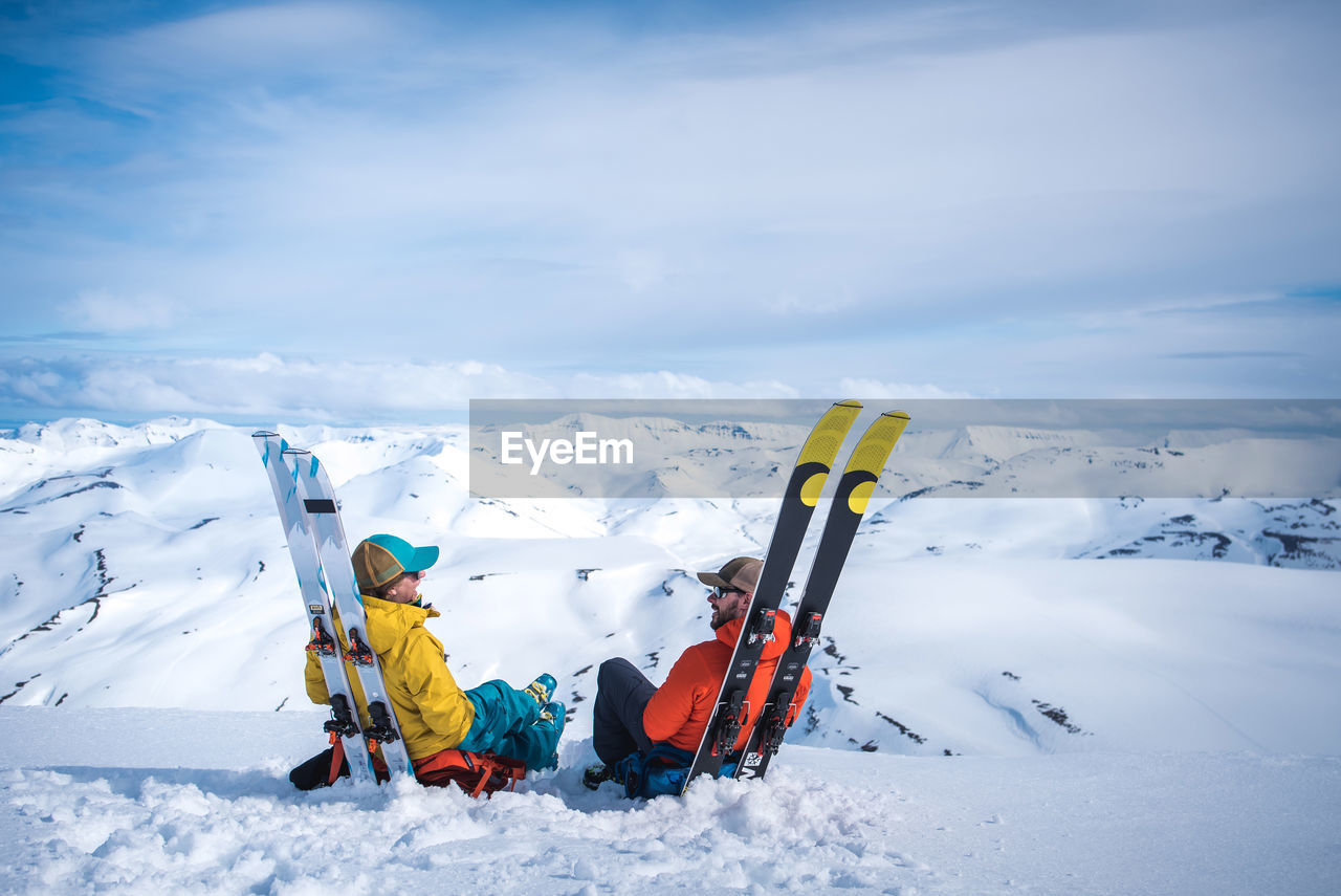 Woman and man sitting against skis with mountains in background