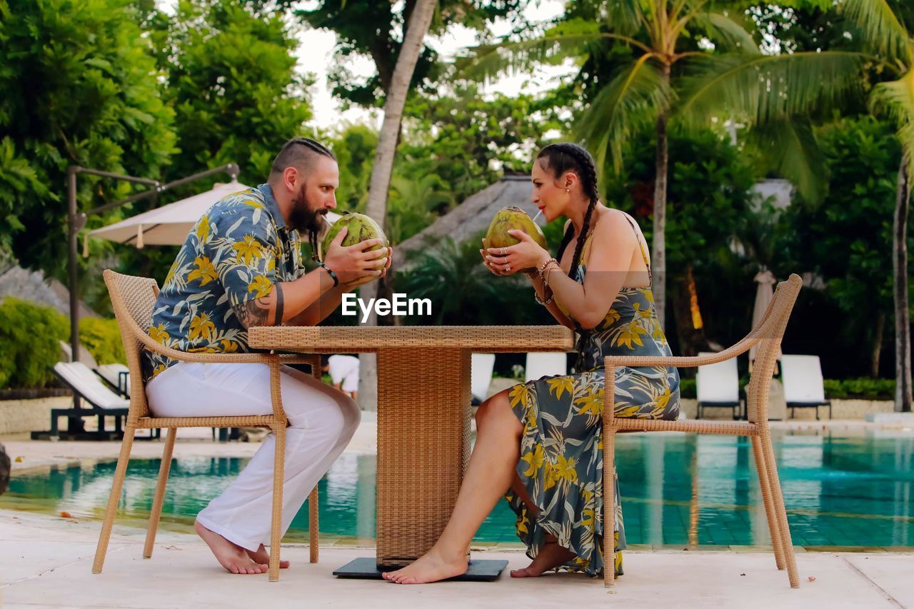 Side view full length of couple drinking coconut water while sitting on chairs at poolside