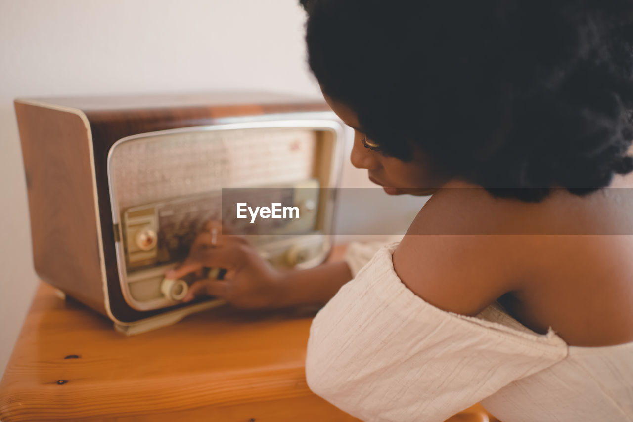 Side view of young african american female sitting at table and tuning old fashioned radio receiver while resting at home