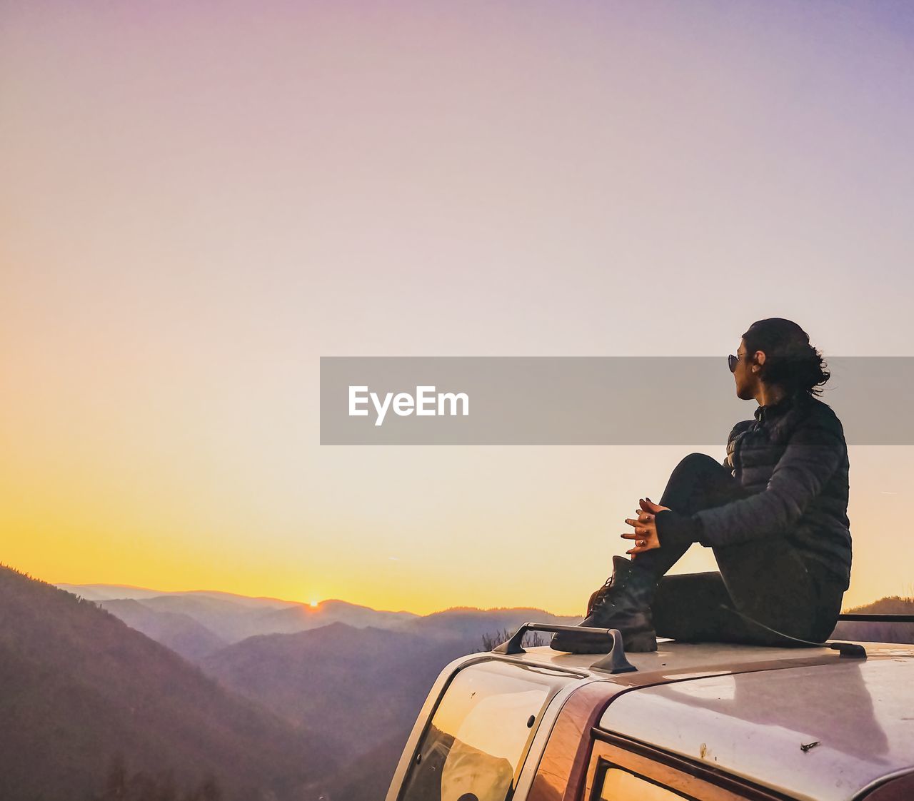 Woman sitting on car against sky during sunset