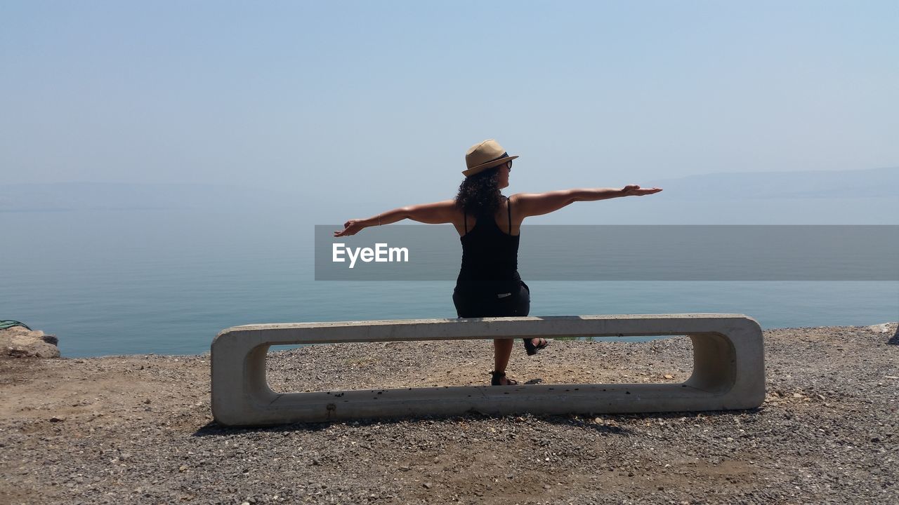 Rear view of woman sitting on concrete seat by lakeshore against sky