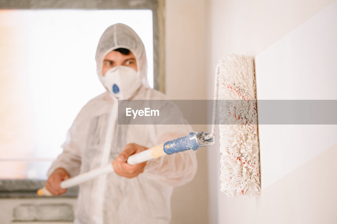 Man in protective workwear working and holding paint roller while standing against wall at home