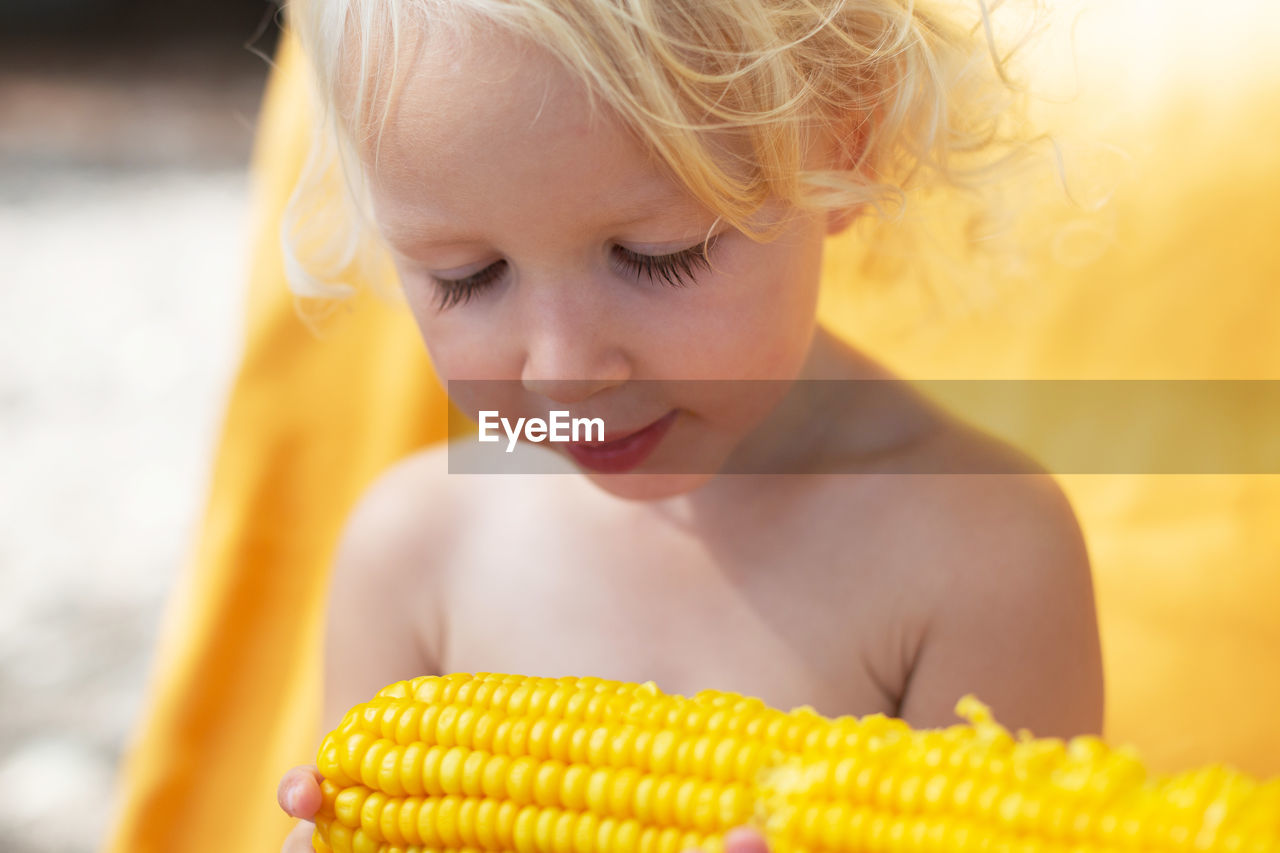 Blond and curly toddler girl eating corn on the beach. yellow colors, summer mood