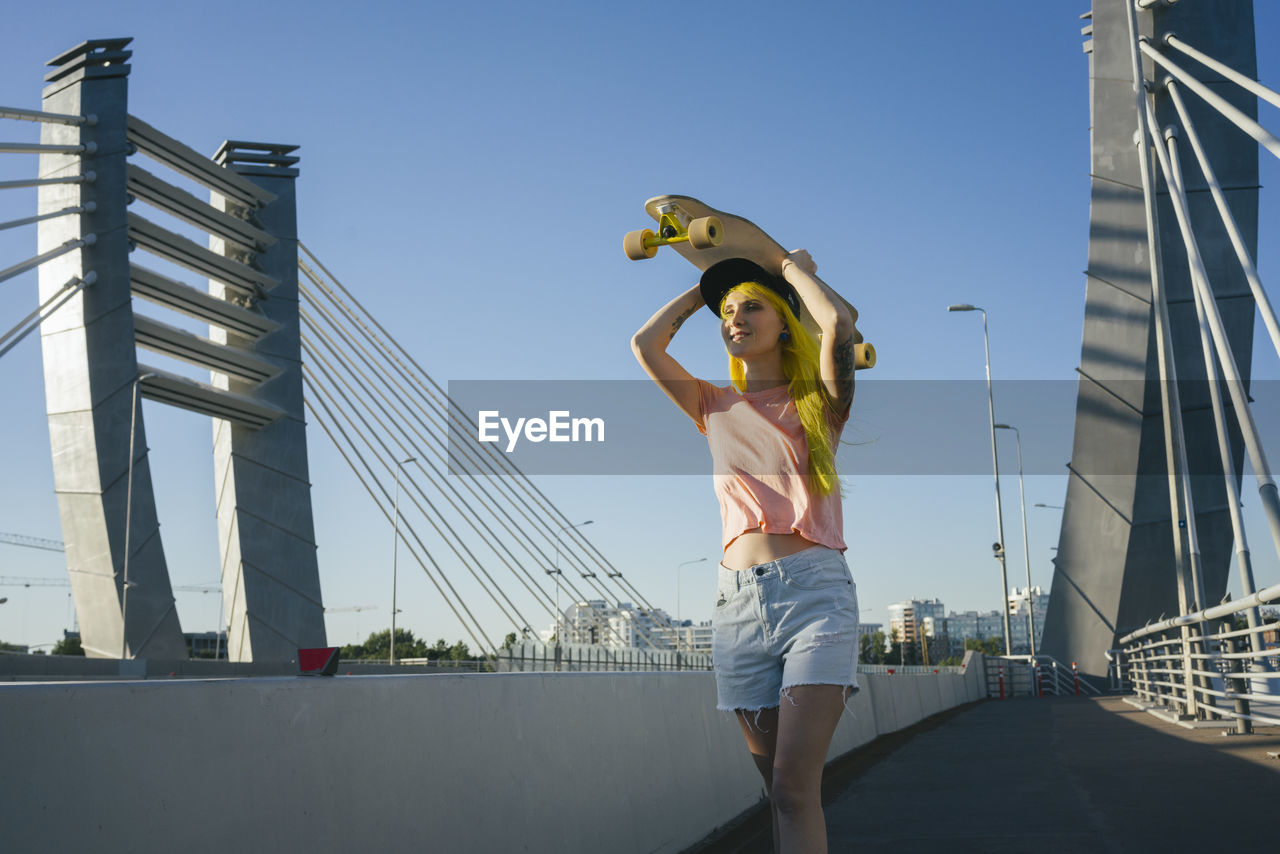 Young woman lifting skateboard on head while standing on bridge during sunny day