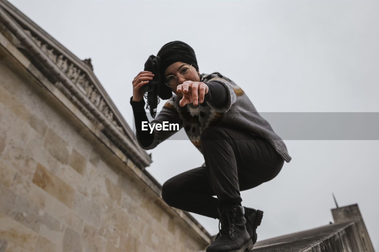 Low angle portrait of woman gesturing while crouching in city against sky