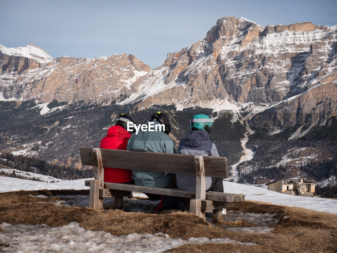 Three skiers relaxing on a wooden bench admiring the mountain panorama in front of them.