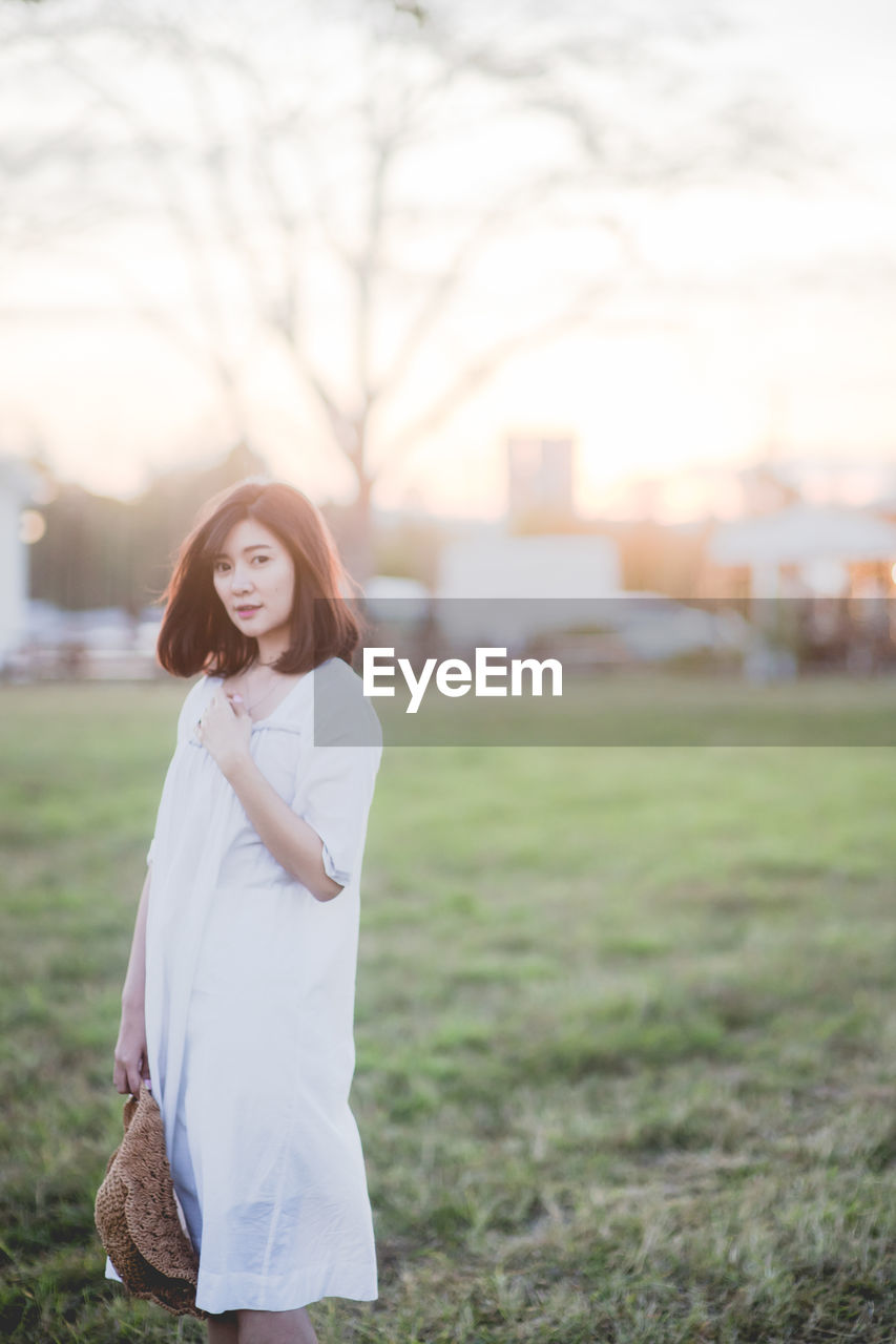 Portrait of beautiful woman holding sun hat while standing on field against sky during sunset