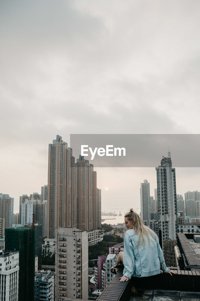 Rear view of woman sitting on building terrace against sky in city