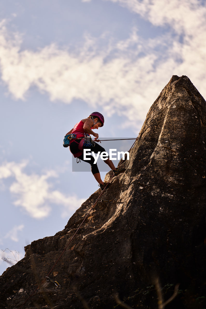Low angle full length of active senior woman climbing on rocky cliff against sky