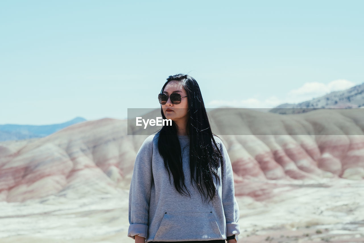 PORTRAIT OF YOUNG WOMAN WEARING SUNGLASSES STANDING IN DESERT